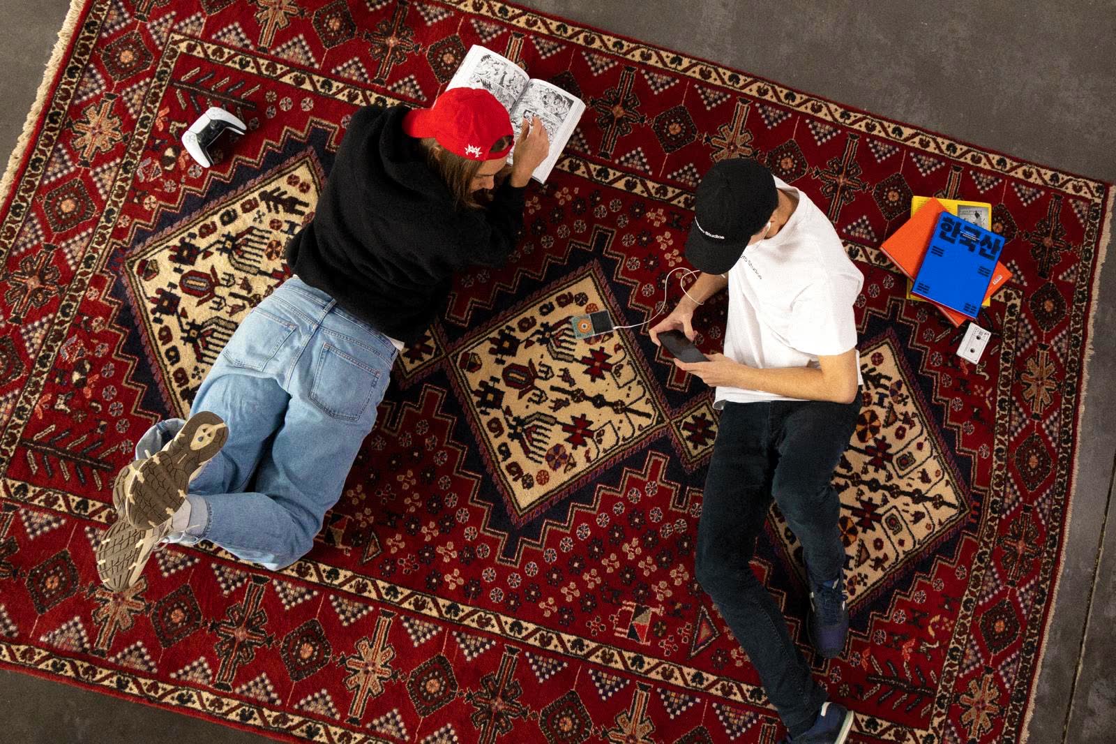 Two friends lying on a red patterned rug, one reading a comic, the other on his phone.